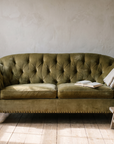 Olive velvet tufted sofa in morning light with a linen throw, an open book and a stool with coffee