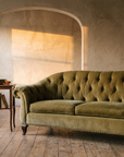 Olive velvet tufted sofa in a plaster salon with amber light through an arch, a side table with books and tea