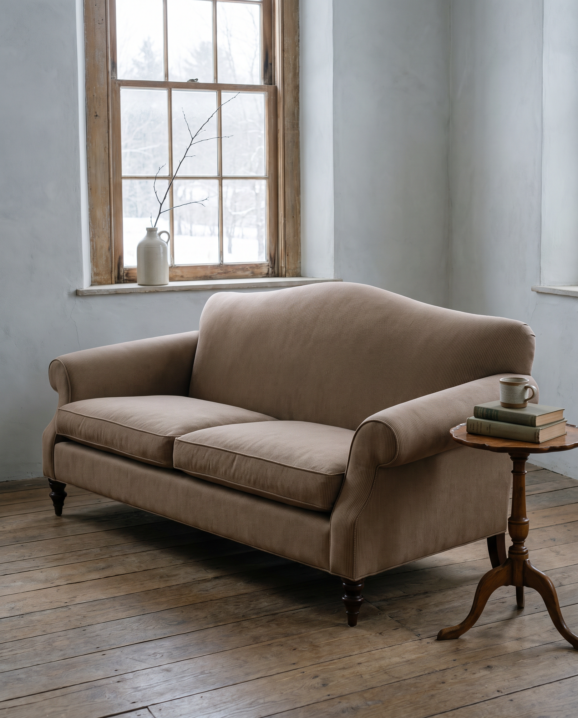 Taupe camelback sofa in a cool grey winter room with a snowy window and a tripod side table