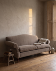 Taupe camelback sofa at dusk beside half-closed timber shutters with a slat of honey light, an open book and a glass of wine