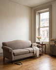 Taupe camelback sofa in a Harlem brownstone parlor beside a tall window with roses on a side table