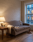 Taupe camelback sofa in a West Village apartment at dusk with a glowing linen lamp and limewashed brick