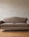 Taupe ticking camelback sofa against a hand-troweled plaster wall with a tall pale stoneware vase of bare branches