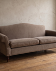 Three-quarter view of the taupe camelback sofa from the right with a weathered stool and stoneware vase by the window