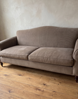 Three-quarter view of the taupe camelback sofa from the left with a stoneware vase of branches in the corner