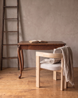The oak writing desk with a pale ash armchair draped in an oatmeal linen blanket and a tall weathered wood ladder leaning on the plaster wall