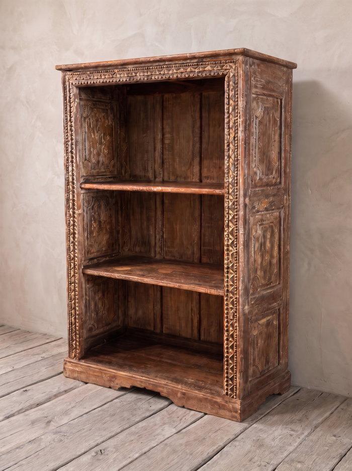 Carved Patina Bookcase, three-quarter view, against greige lime plaster on a wide bleached oak floor.