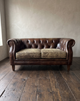 Vintage tobacco leather chesterfield loveseat, front view, greige plaster wall and dark reclaimed wood floor
