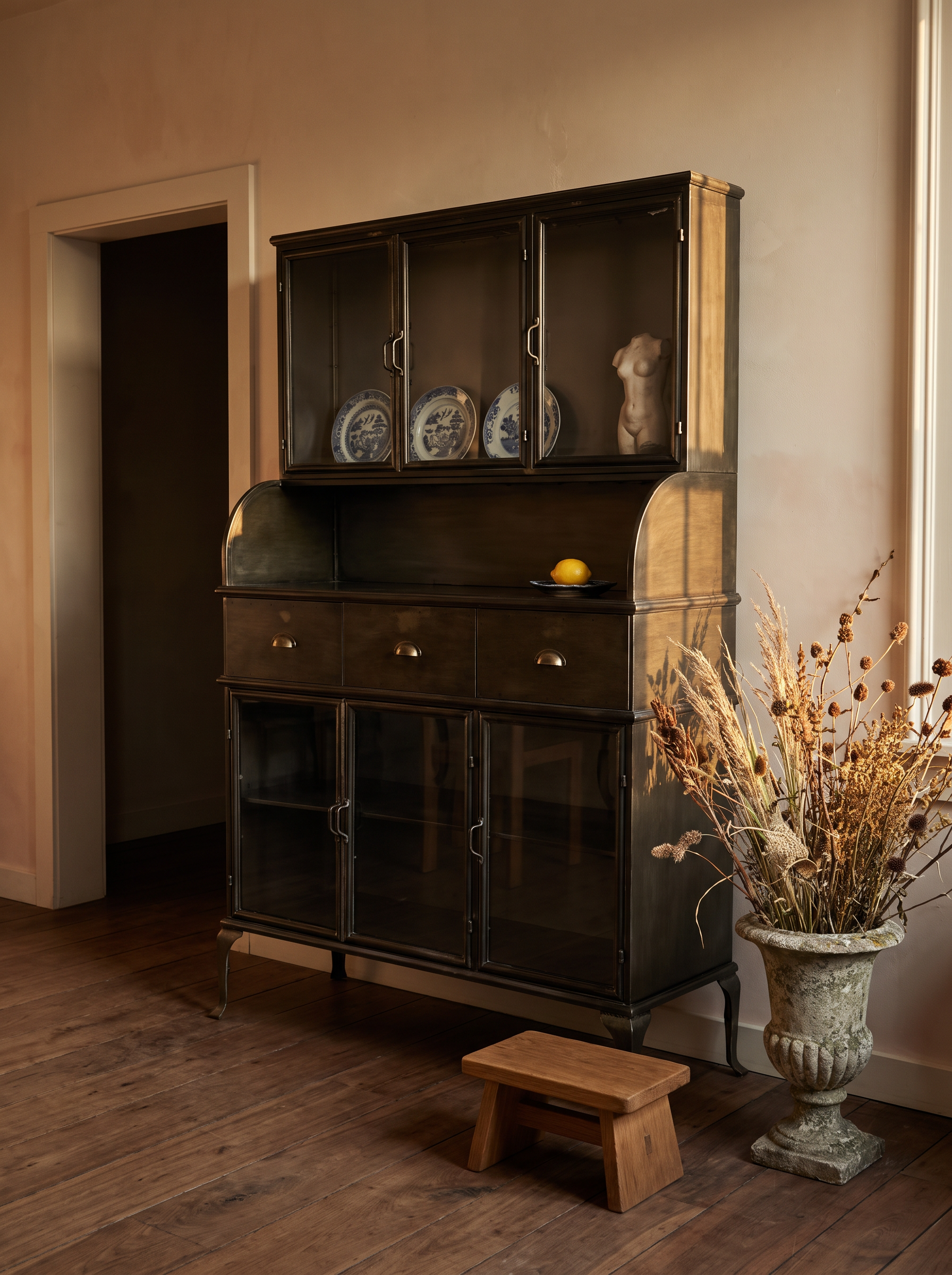 Steel hutch in a warm golden-lit room with blue and white plates behind the glass, an oak stool and a stone urn of dried grasses