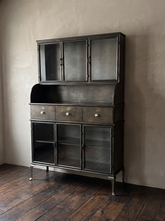 Apothecary steel hutch with glass doors and brass cup pulls against greige plaster on a dark antique oak floor, three-quarter view