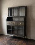 Apothecary steel hutch with glass doors and brass cup pulls against greige plaster on a dark antique oak floor, three-quarter view