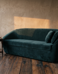 Green velvet curved sofa beside an antique side table with old books and tea, late sun across a plaster wall