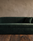 Front view of a long deep green velvet curved sofa with two ruched velvet pillows in a plaster room