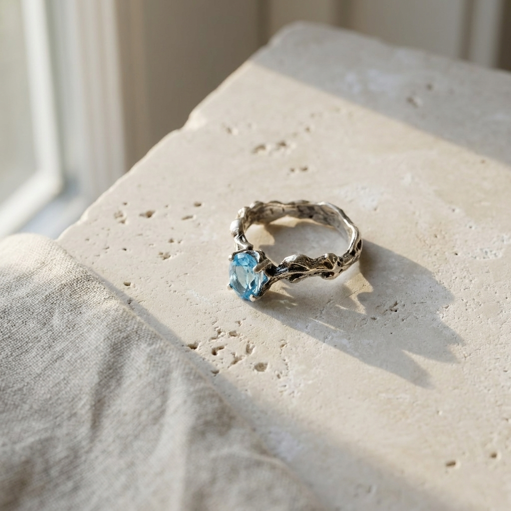 Sterling silver twig ring set with an oval blue topaz, laid flat on pale stone