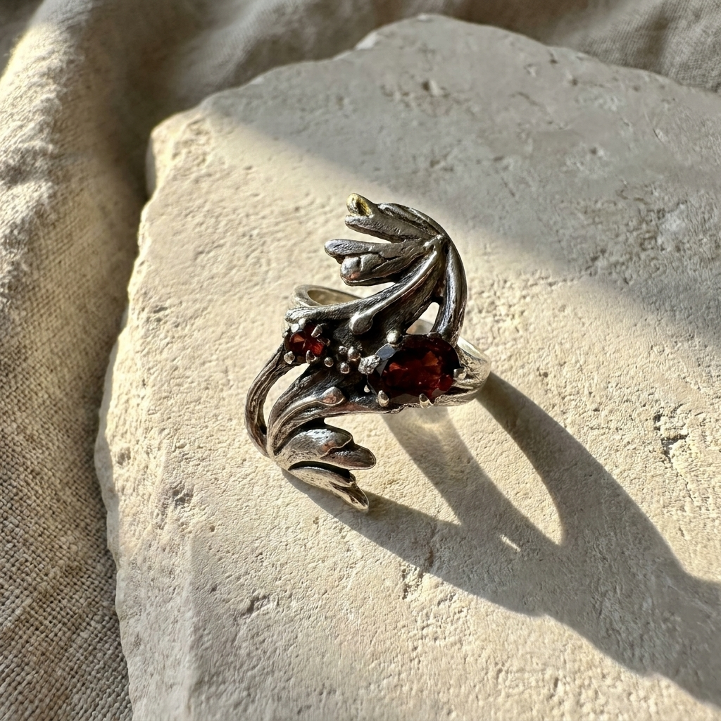 Sculptural sterling silver leaf ring set with two deep red garnets, laid flat on pale stone