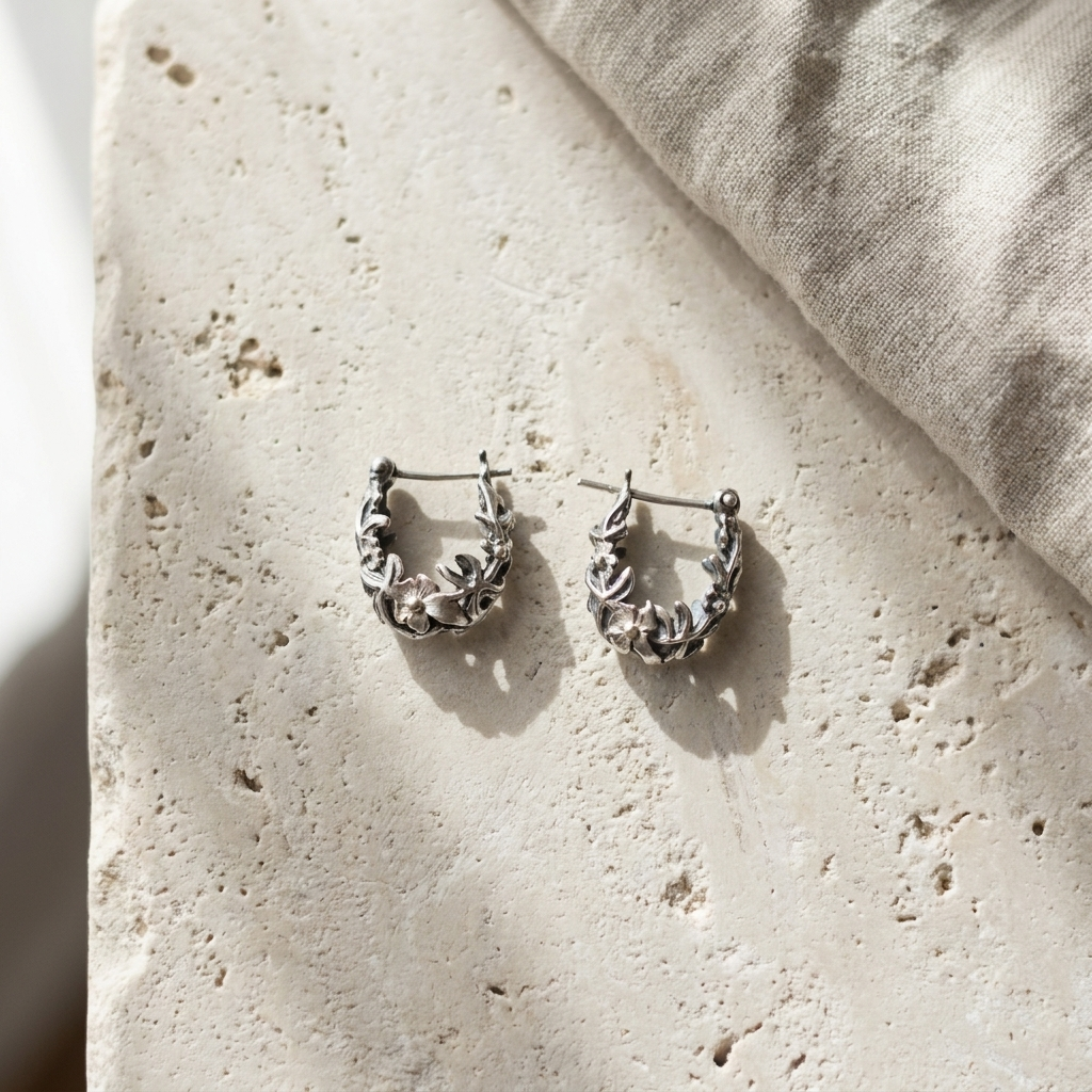 Sterling silver floral relief huggie hoop earrings, laid flat on pale stone