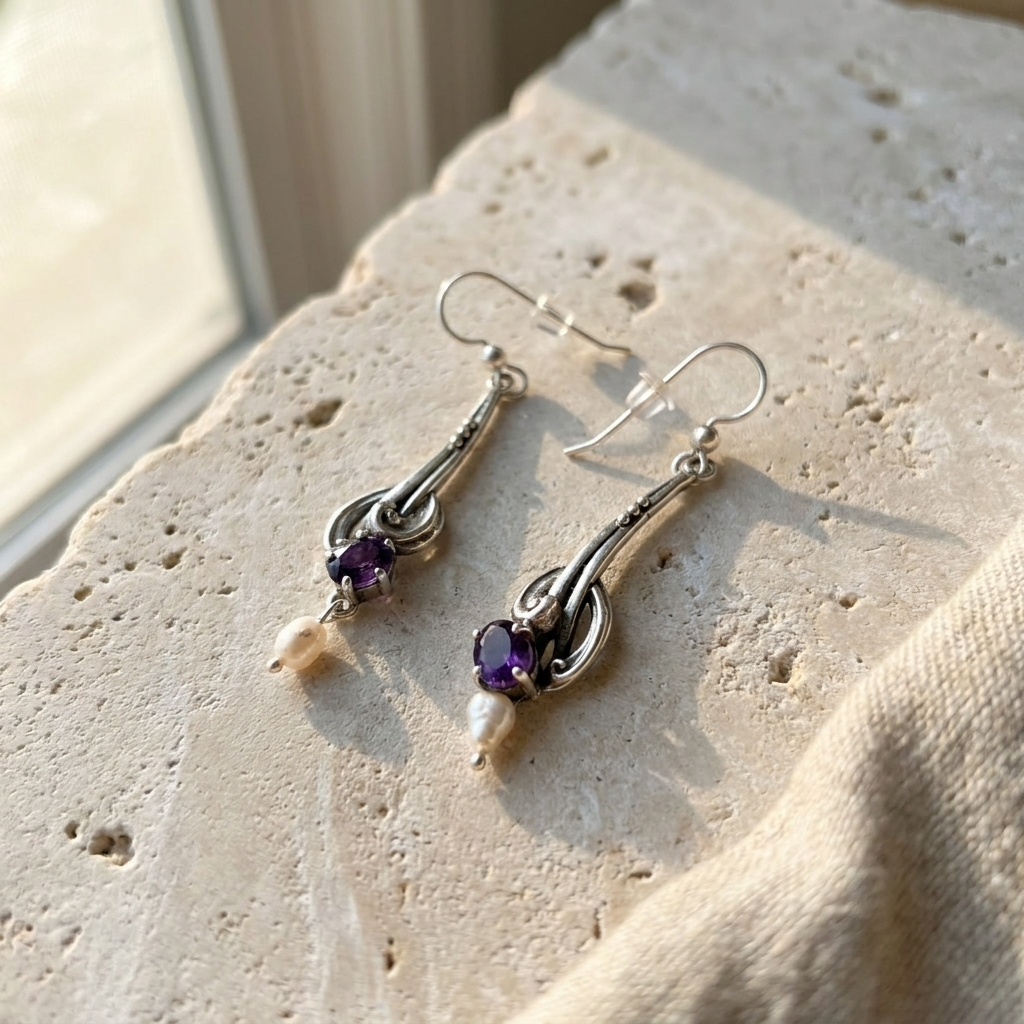 Sterling silver drop earrings with amethyst and baroque pearls, laid flat on pale stone