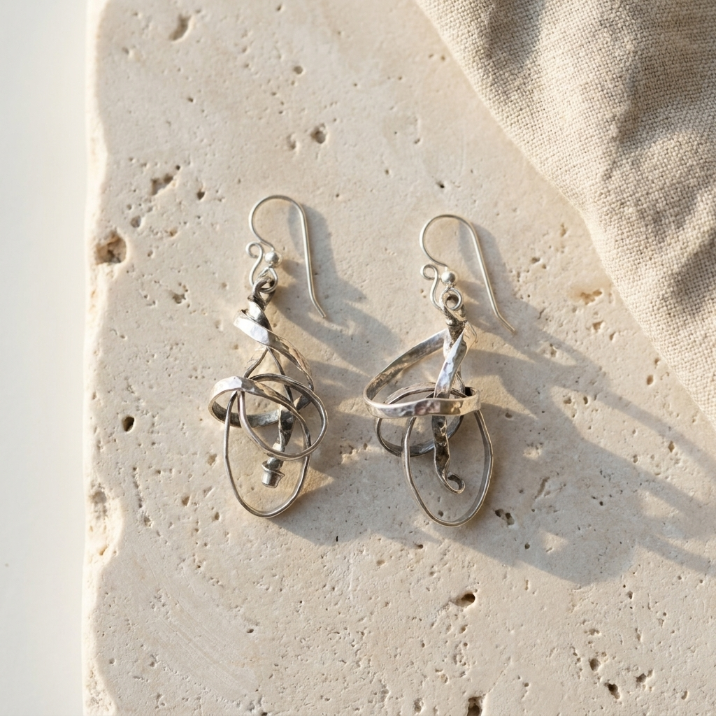 Twisted hammered sterling silver ribbon earrings with inner hanging loops, laid flat on pale stone
