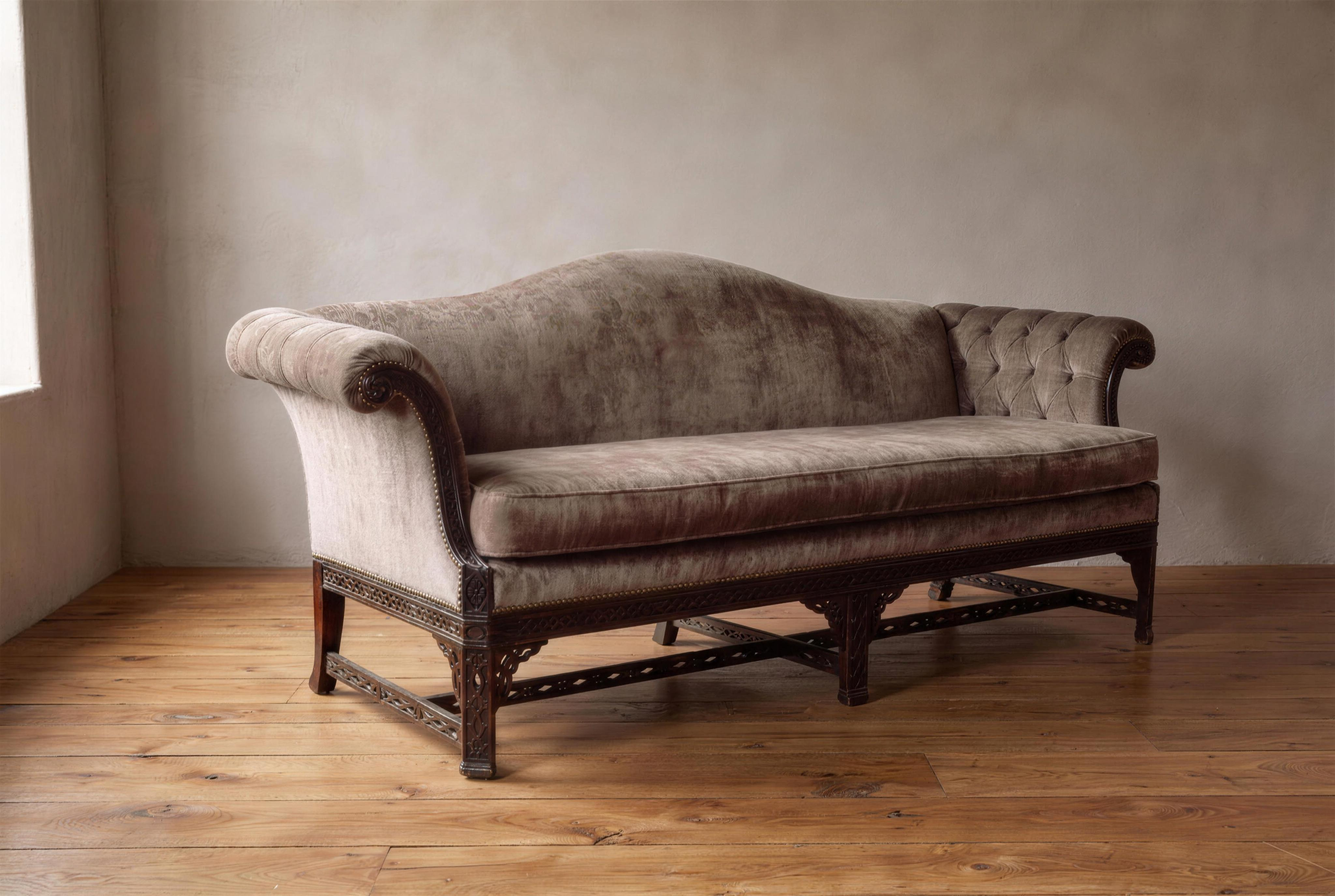The Dorian, an antique carved Chinese Chippendale camelback sofa in taupe velvet, front view, against a plaster wall on a reclaimed wood floor