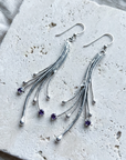 Long sterling silver comet earrings with fanned rods and small amethysts, laid flat on pale stone