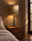 Mirrored column table lamp illuminated beside a bed in a plaster room