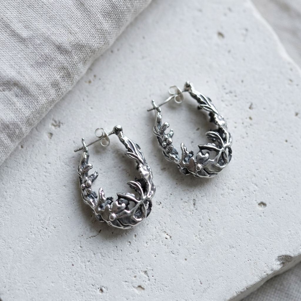 Cast sterling silver branch and leaf oval hoop earrings laid flat on pale stone