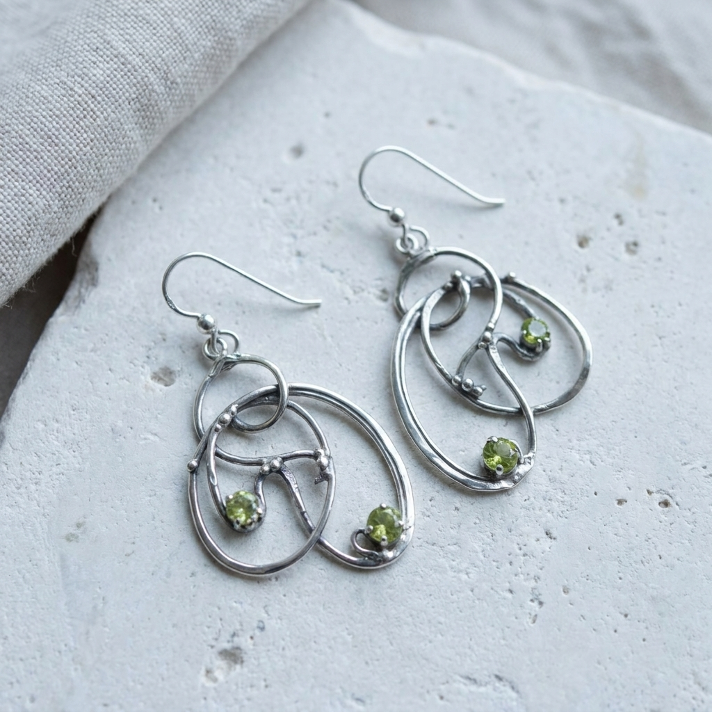 Open looping sterling silver Art Nouveau earrings set with peridots, laid flat on pale stone