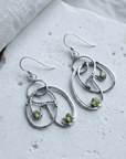 Open looping sterling silver Art Nouveau earrings set with peridots, laid flat on pale stone
